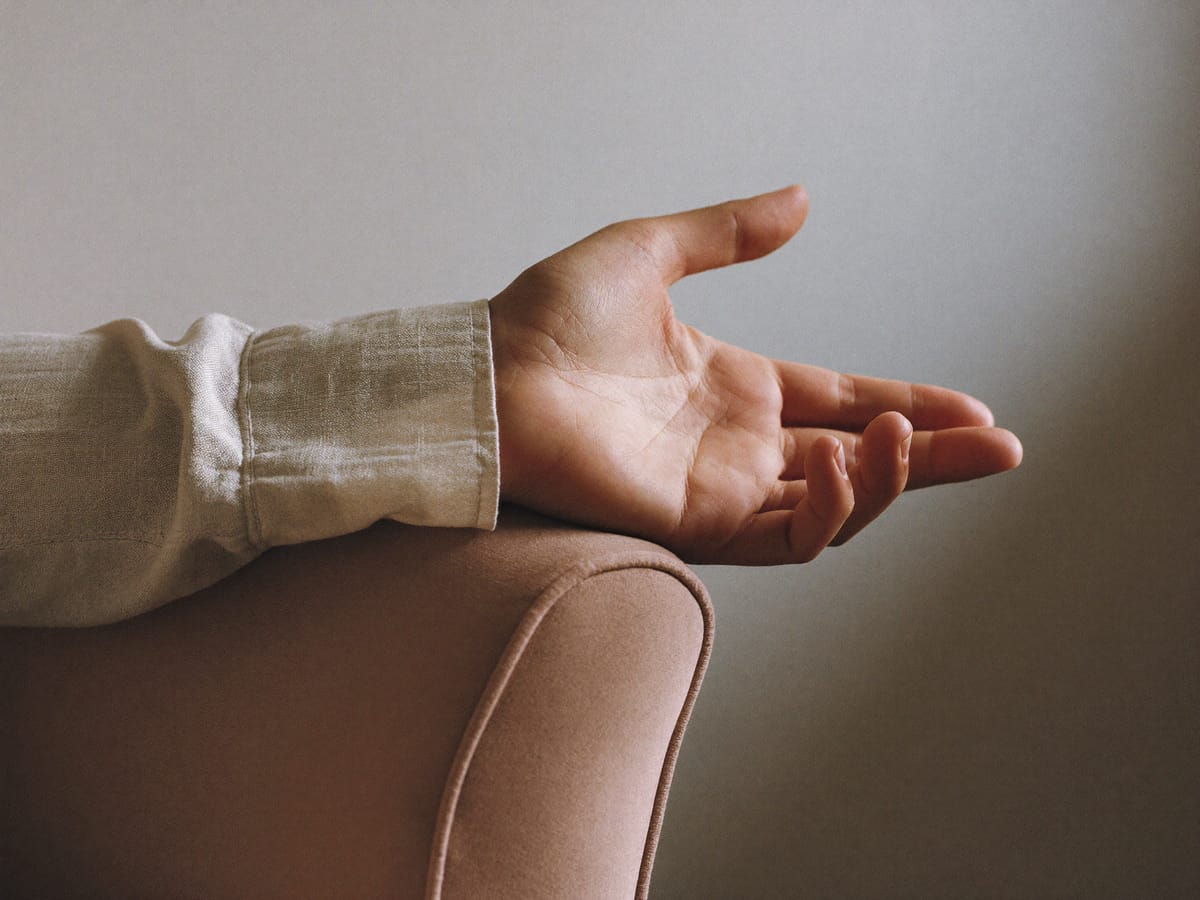One hand at rest, palm up, on the upholstered arm of a chair, in natural light against a plain wall.